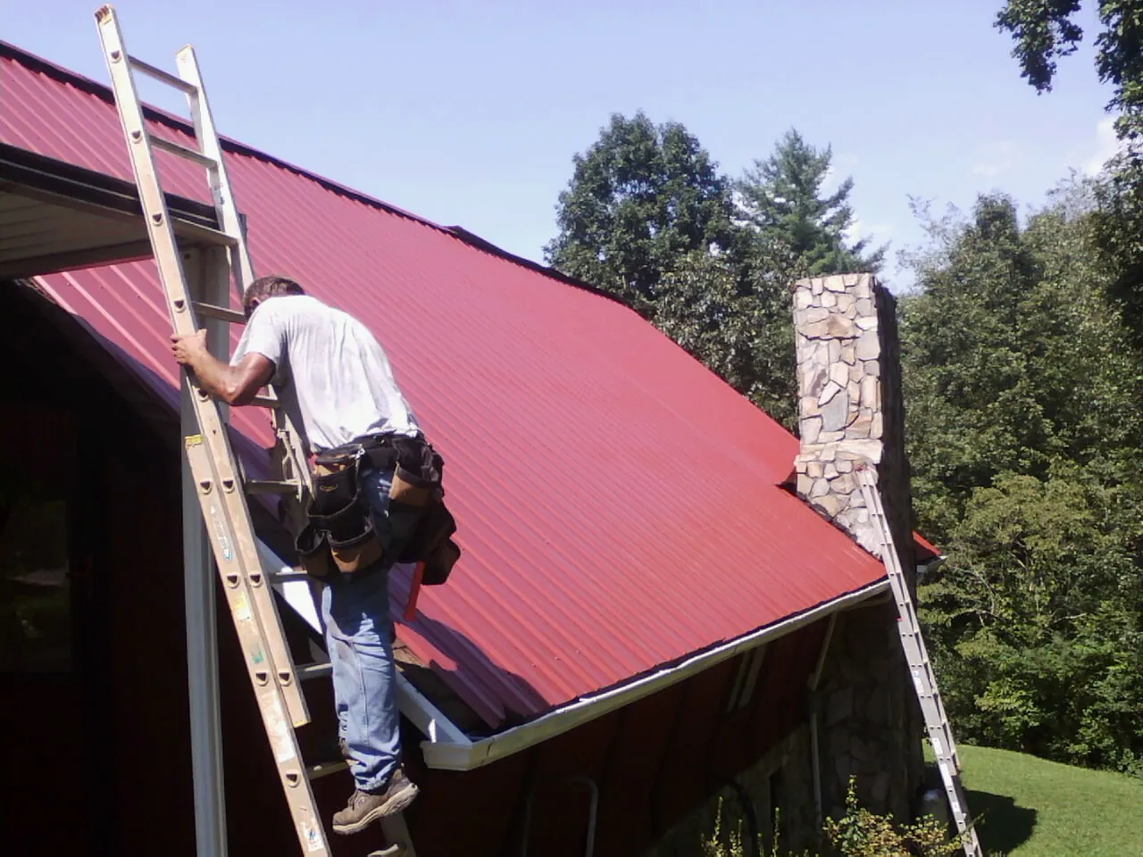 Roofer on a ladder beside a red metal roof and stone chimney — Soffit & Fascia Repair by Roofing Brothers in Waterloo